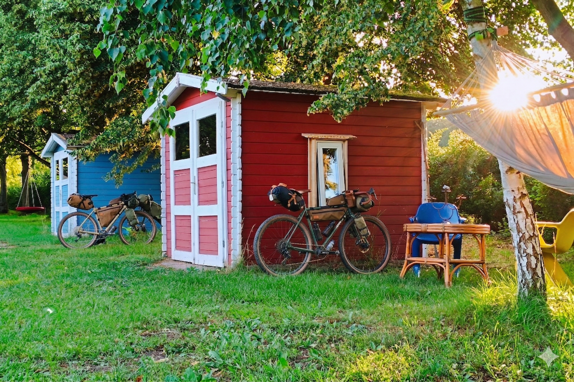 Ein Foto im warmen, goldenen Licht des Sonnenuntergangs zeigt wei kleine, rot gestrichene Holzhütten mit weißen Details. An der linken Hütte lehnt ein Tourenrad mit Gepäcktaschen. Rechts bricht das Sonnenlicht durch die Blätter eines Baumes, neben dem eine Hängematte an einer Birke hängt. Die gesamte Szene ist von Bäumen umgeben.