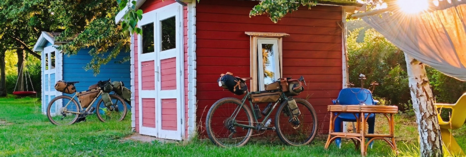 Ein Foto im warmen, goldenen Licht des Sonnenuntergangs zeigt wei kleine, rot gestrichene Holzhütten mit weißen Details. An der linken Hütte lehnt ein Tourenrad mit Gepäcktaschen. Rechts bricht das Sonnenlicht durch die Blätter eines Baumes, neben dem eine Hängematte an einer Birke hängt. Die gesamte Szene ist von Bäumen umgeben.